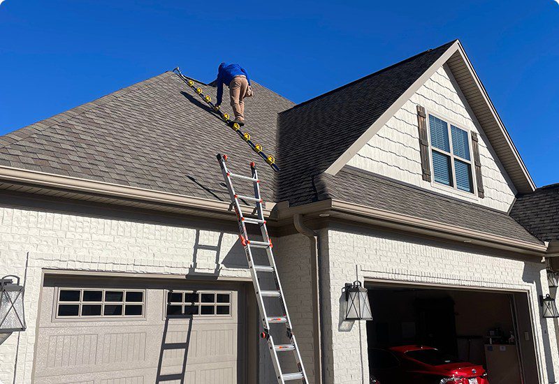Roofer installing shingles on a steep residential roof using safety equipment and a ladder.